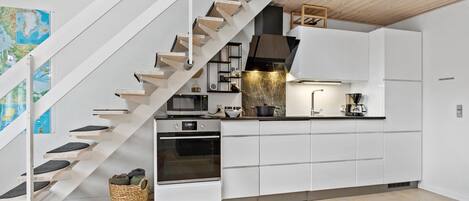 A modern kitchen with white cabinetry, a built-in oven, a microwave, and a coffee maker. There is a wooden staircase with a basket of shoes at the bottom.