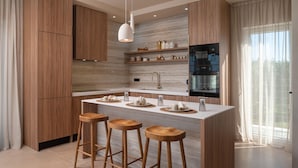 Eine moderne Küche mit Holzausstattung, einer zentralen Kochinsel mit Barhocker, einem eingebauten Backofen und einem großen Fenster mit durchsichtigen Vorhängen.