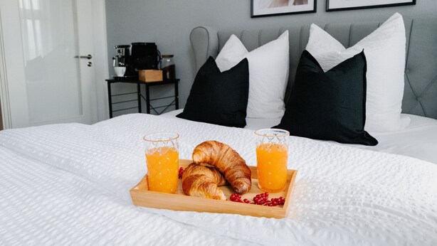 En snyggt bäddad säng med vita sängkläder, ett bricka med croissanter och apelsinjuice och en kaffemaskin på ett sidobord.