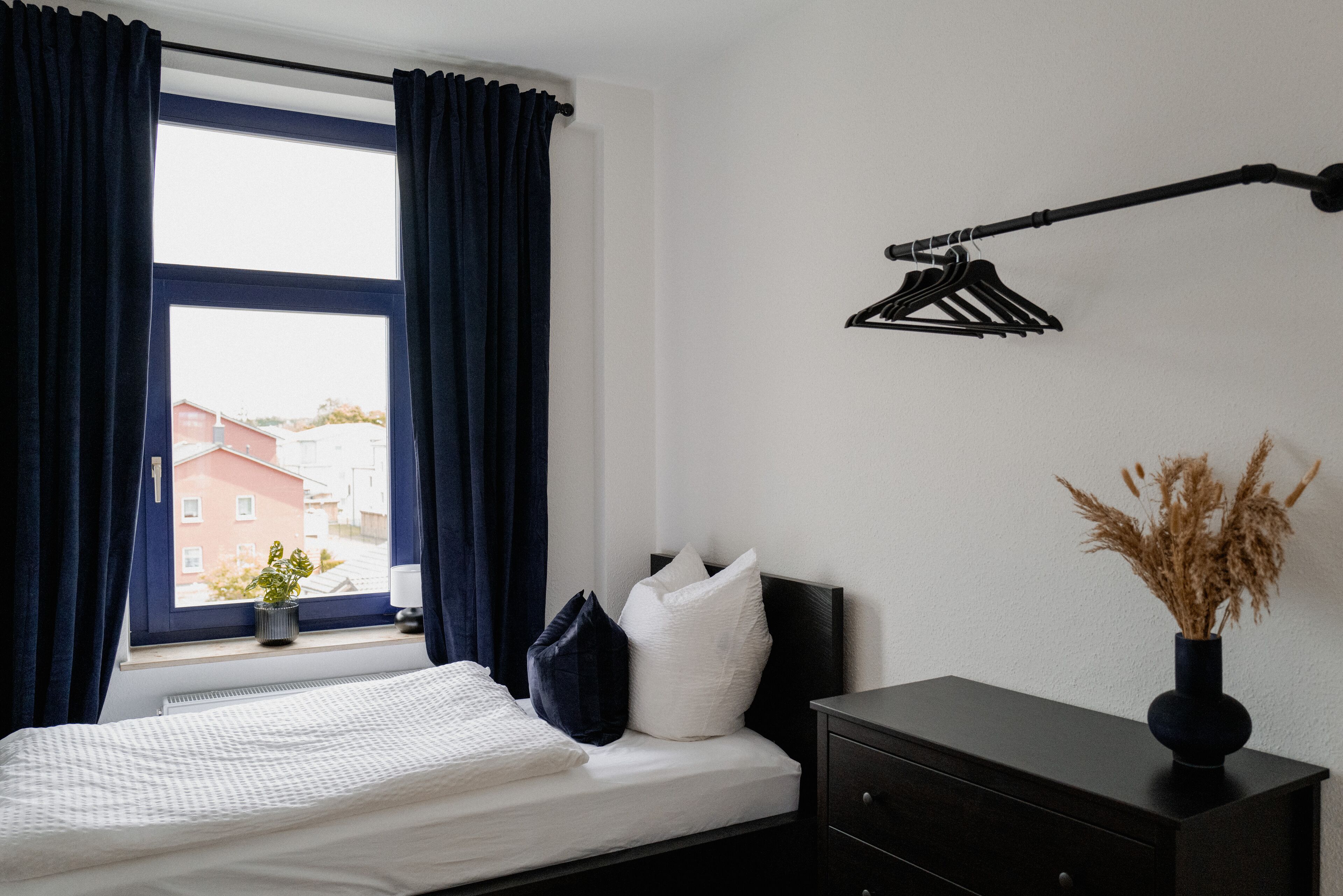 A bedroom with a bed, a window with curtains, a bedside table with a vase, and a wall-mounted light fixture.