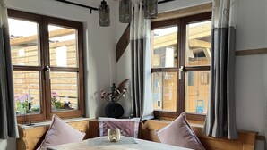 Un coin repas en bois avec une table et des bancs, de grandes fenêtres avec des rideaux et des lampes suspendues.