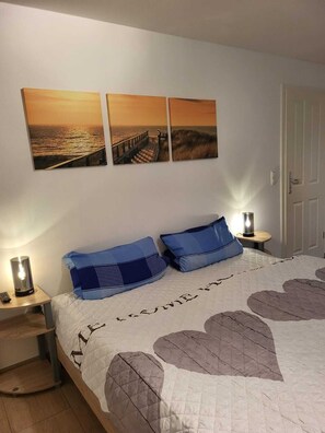 A bedroom with a bed, two wall-mounted pictures, a nightstand with a lamp, and a door. - Ferienwohnung Lanckensburg 1 (Altenkirchen)