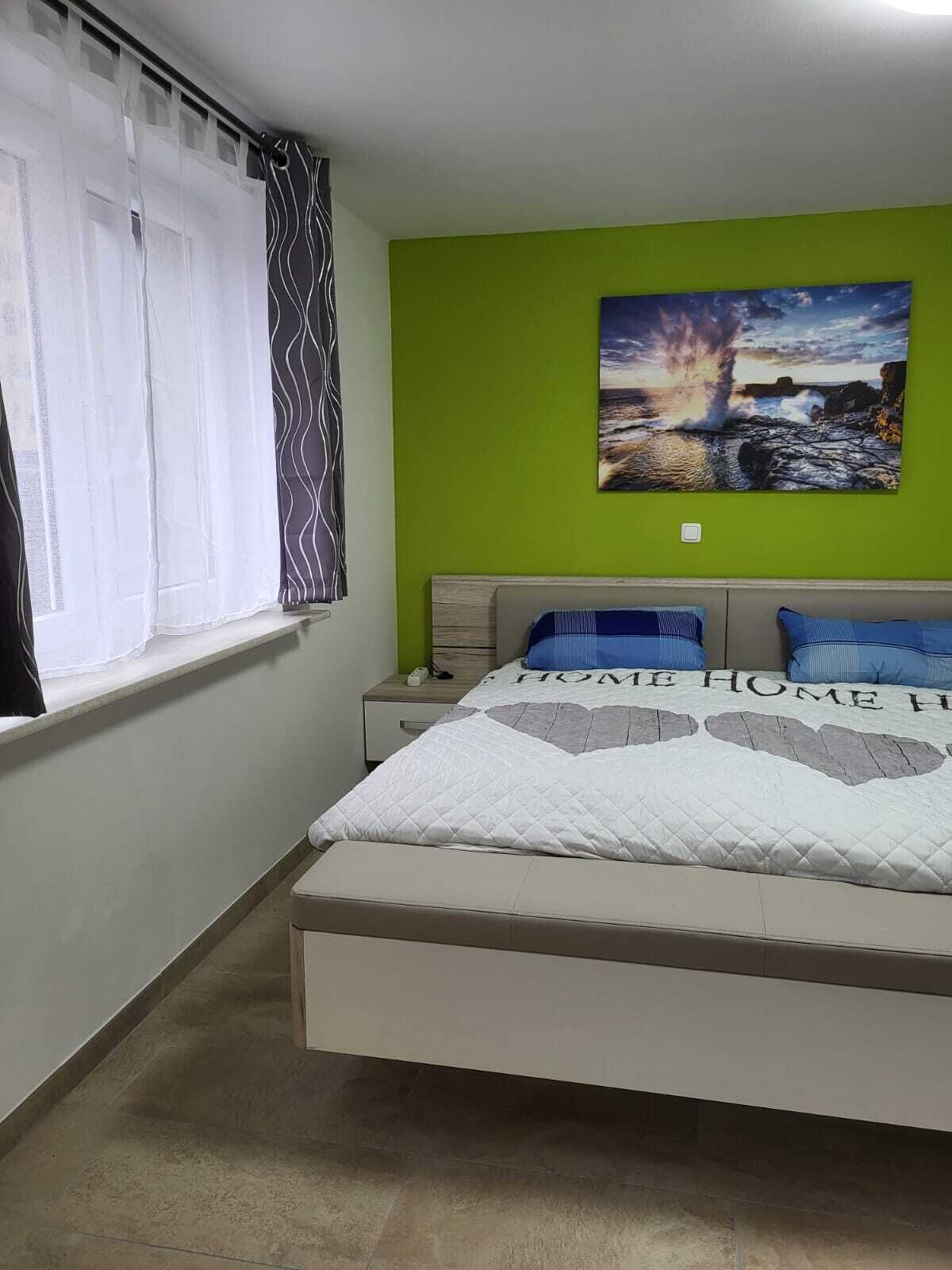 Un dormitorio con cama, mesita de noche, pared verde y ventana con cortinas.