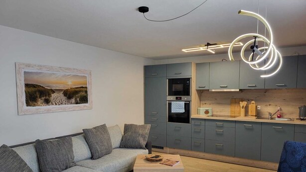 Un salon moderne avec un canapé gris, une table ronde en bois et une cuisine dotée de meubles gris et d’un four encastré.