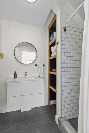 A bathroom with a white vanity, a round mirror, a wooden shelf with towels, and a shower area with a white curtain. - Queens Suite 1BR Free Parking (St. John's)
