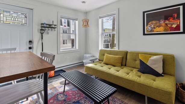 A modern living room with a sofa, a dining table, and a bench. There are windows, a hanging light fixture, and a framed picture on the wall. - Queens Suite 1BR Free Parking (St. John's)