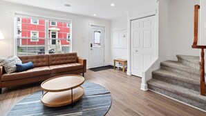 A modern living room with a brown leather sofa, a round wooden coffee table, and a view of a red building through large windows. - Wood House King Bd 2 Queens Fire Pit (St. John's)