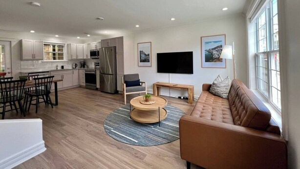 A modern living room with a brown sofa, a coffee table, and a TV mounted on the wall. - Wood House King Bd 2 Queens Fire Pit (St. John's)