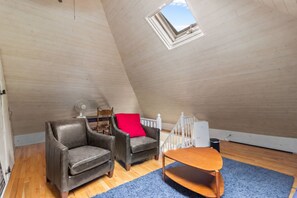 A loft bedroom with a sloped ceiling, a skylight, a wooden floor, two leather armchairs, a small wooden table, and a white railing.