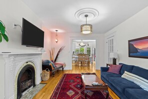 A living room with a fireplace, television, blue sofa, and a dining area with chairs and a chandelier.