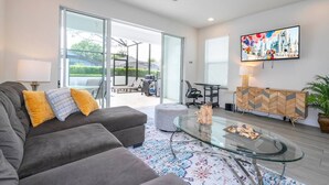A modern living room with a grey sofa, a glass coffee table, and a television mounted on the wall. - Comfortable home Near Disney & Private Pool 17414 (Clermont)