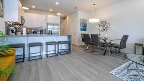 A modern kitchen with a central island, bar stools, and a dining area with a glass table and chairs. - Comfortable home Near Disney & Private Pool 17414 (Clermont)