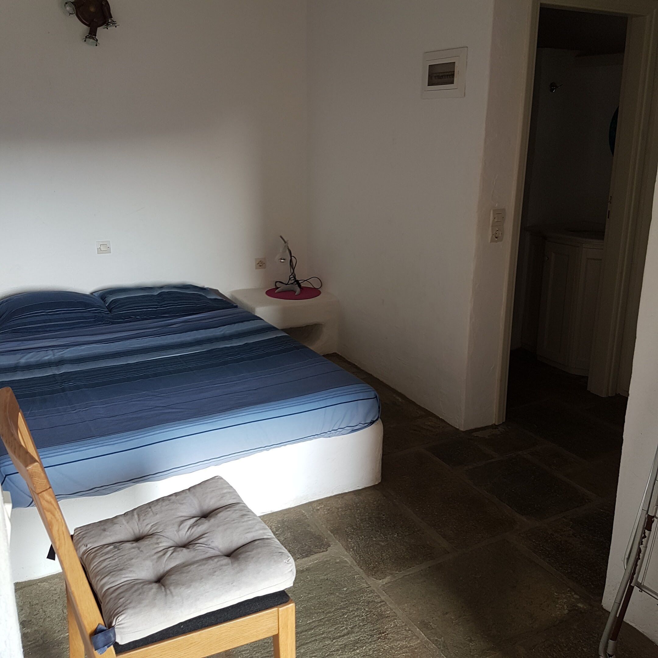 Een slaapkamer met een bed, een nachtkastje met lamp, een stoel en een deur die naar een andere kamer leidt.