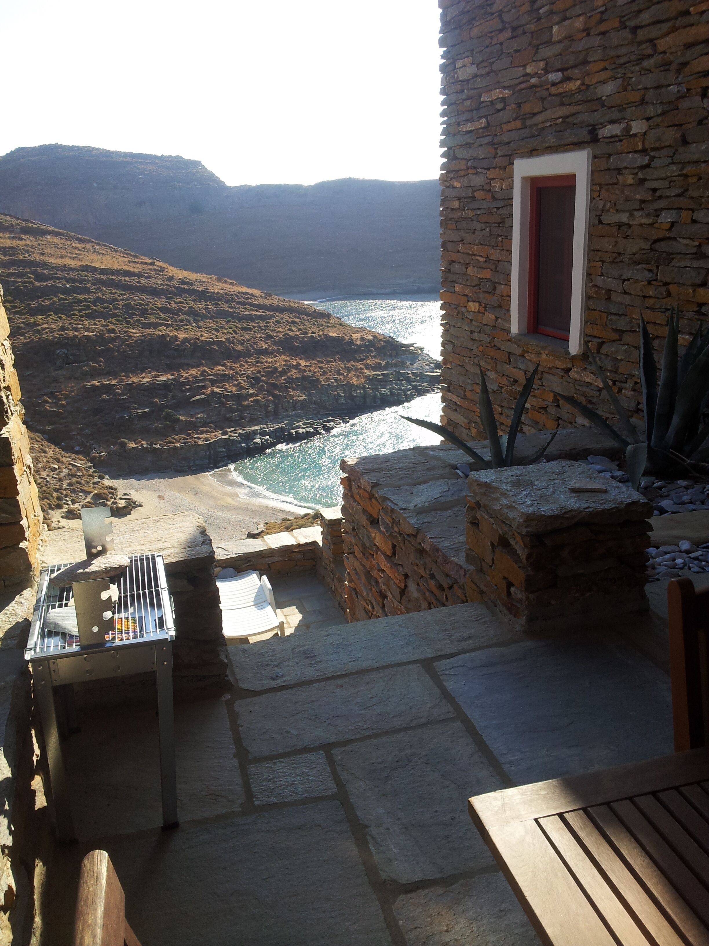 A stone patio with a table and chairs, overlooking a coastal area with a view of the sea and distant hills.