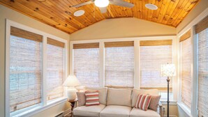 Ein gemütliches Wohnzimmer mit Holzbalkendecke, großen Fenstern mit Jalousien, einem beige gefärbten Sofa mit gestreiften Kissen und einer Stehlampe.