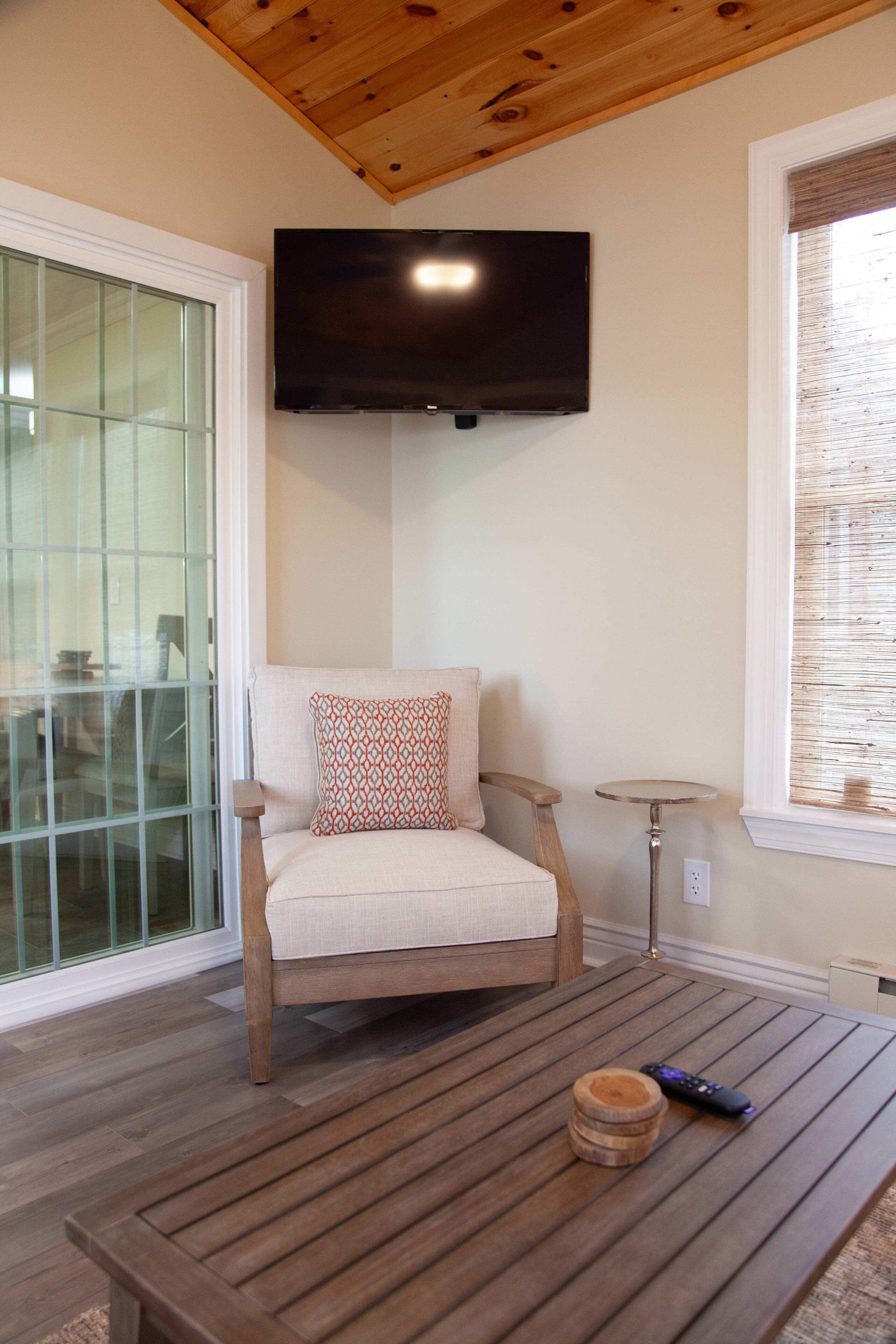 Une pièce chaleureuse avec un plafond en bois, un téléviseur fixé au mur, un fauteuil confortable, une petite table ronde et une fenêtre avec des stores.
