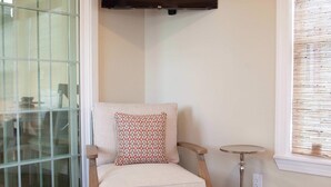 Ein gemütliches Zimmer mit Holzbalkendecke, einem an der Wand befestigten Fernseher, einem gepolsterten Sessel, einem kleinen runden Tisch und einem Fenster mit Jalousien.