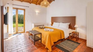 A bedroom with a wooden ceiling, a bed with a yellow blanket, a bedside table, a lamp, and a view of the outdoors through a glass door.