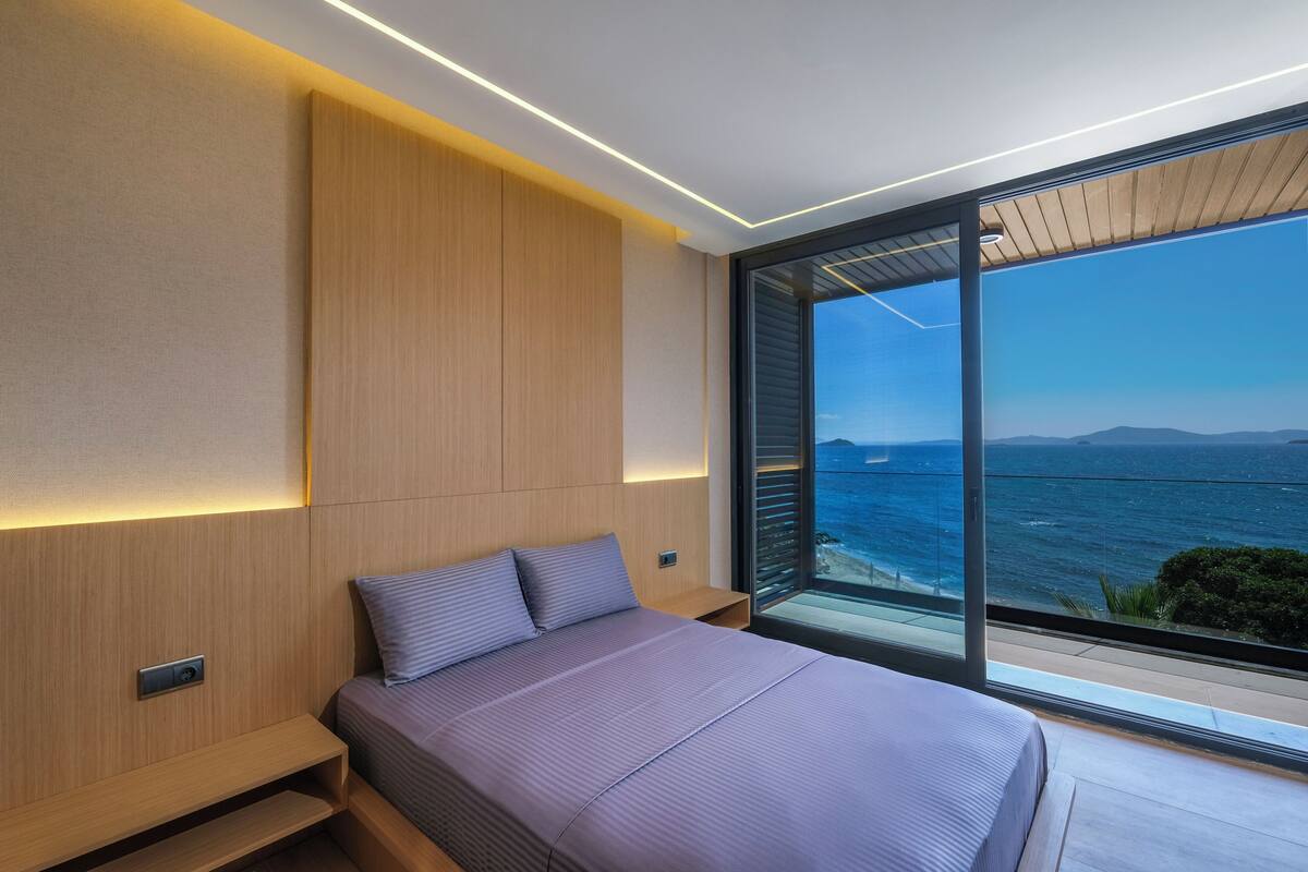 A modern bedroom with a large bed, wooden paneling, a nightstand, and a view of the sea.