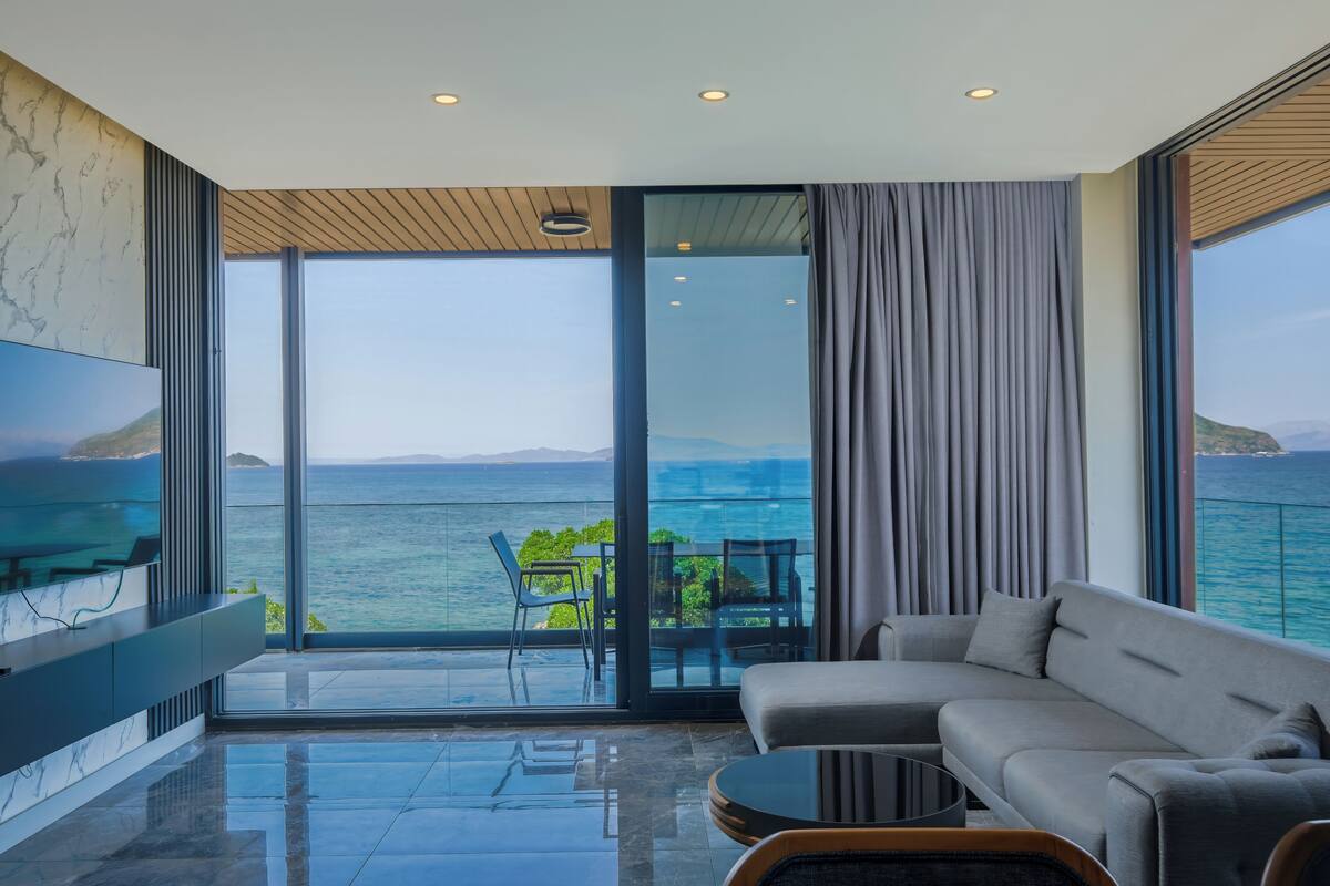 A modern living room with a large flat-screen TV, a sofa, a coffee table, and a view of the sea through floor-to-ceiling windows.
