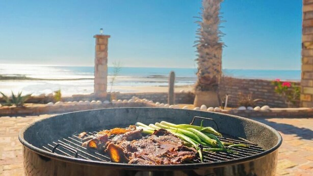 Una parrilla con carne y verduras, frente a una playa y palmeras.