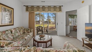 Una sala de estar con un sofá, una mesa de centro y un televisor. Hay una puerta corrediza de vidrio que da a un espacio al aire libre con sillas y una mesa.