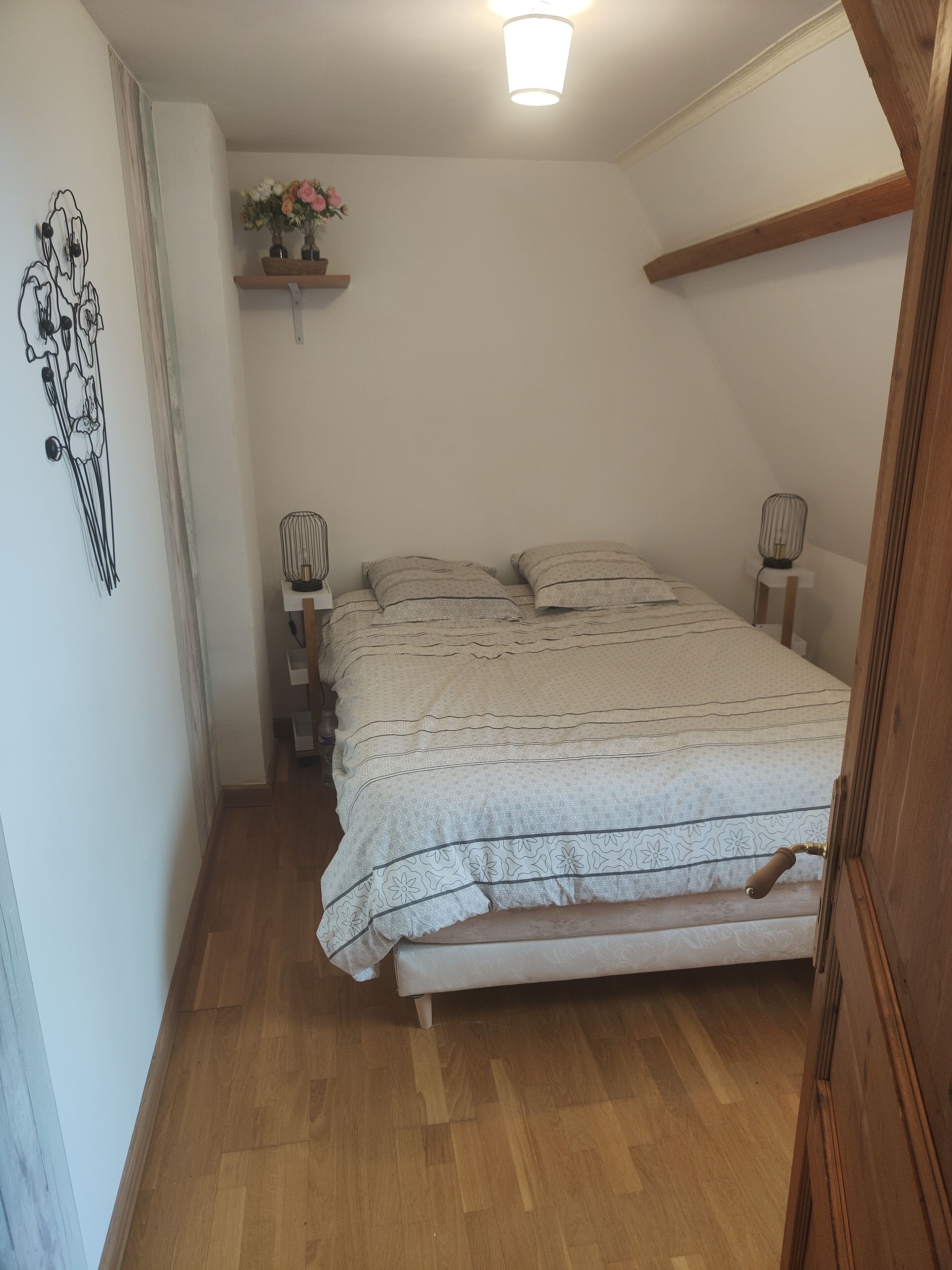 Une chambre avec un plancher en bois, un lit avec une literie à motifs, une table de chevet avec une lampe et une étagère murale décorée de fleurs.