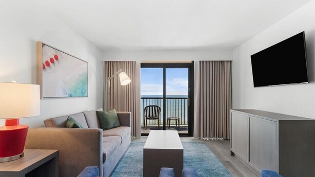 A modern hotel room with a sofa, a coffee table, a TV, and a balcony with a view of the sea. - Oceanfront 3BR Condo: Comfort & Views (Myrtle Beach)