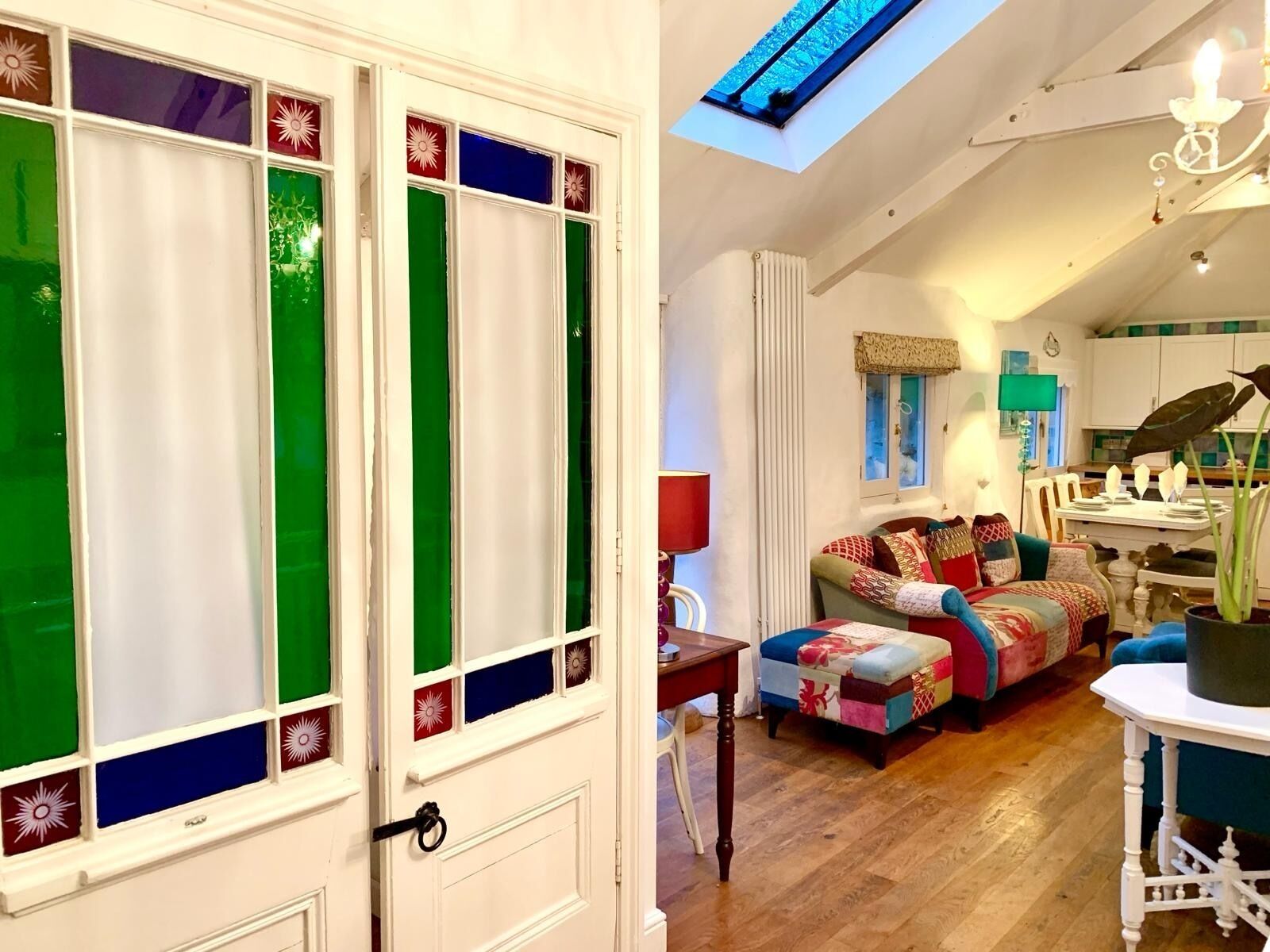 A cozy living room with a sofa, a table, and a chair. There is a window with colorful panes and a skylight above.