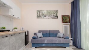 A modern living room with a grey sofa, a kitchen area with grey cabinets, a radiator, and a painting on the wall. - Comfortable Studio | Next to the Royal Baths (Warszawa)