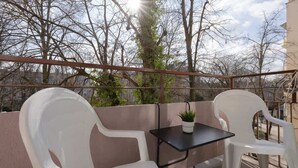 A balcony with two white plastic chairs and a small table, a potted plant, and a view of leafless trees and a building in the background. - Comfortable Studio | Next to the Royal Baths (Warszawa)