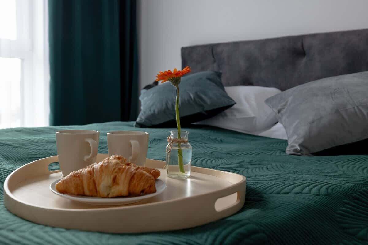 Sänky, jossa on vihreä sängynpeite, tarjotinalus, jossa on croissant, kaksi kuppia ja maljakko, jossa on kukka.