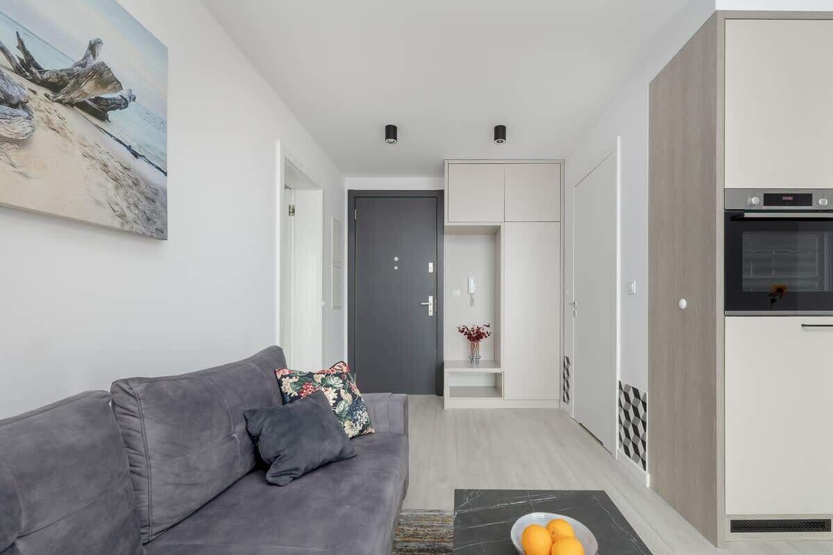 A modern living room with a grey sofa, a coffee table with oranges, and a kitchen area with a built-in oven.