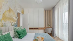 A bedroom with a bed, green pillows, a wooden bedside table with a tray, a desk, and a view of the city through sheer curtains.
