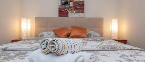 A neatly made bed with a patterned bedspread, two pillows, and rolled towels on the bed.