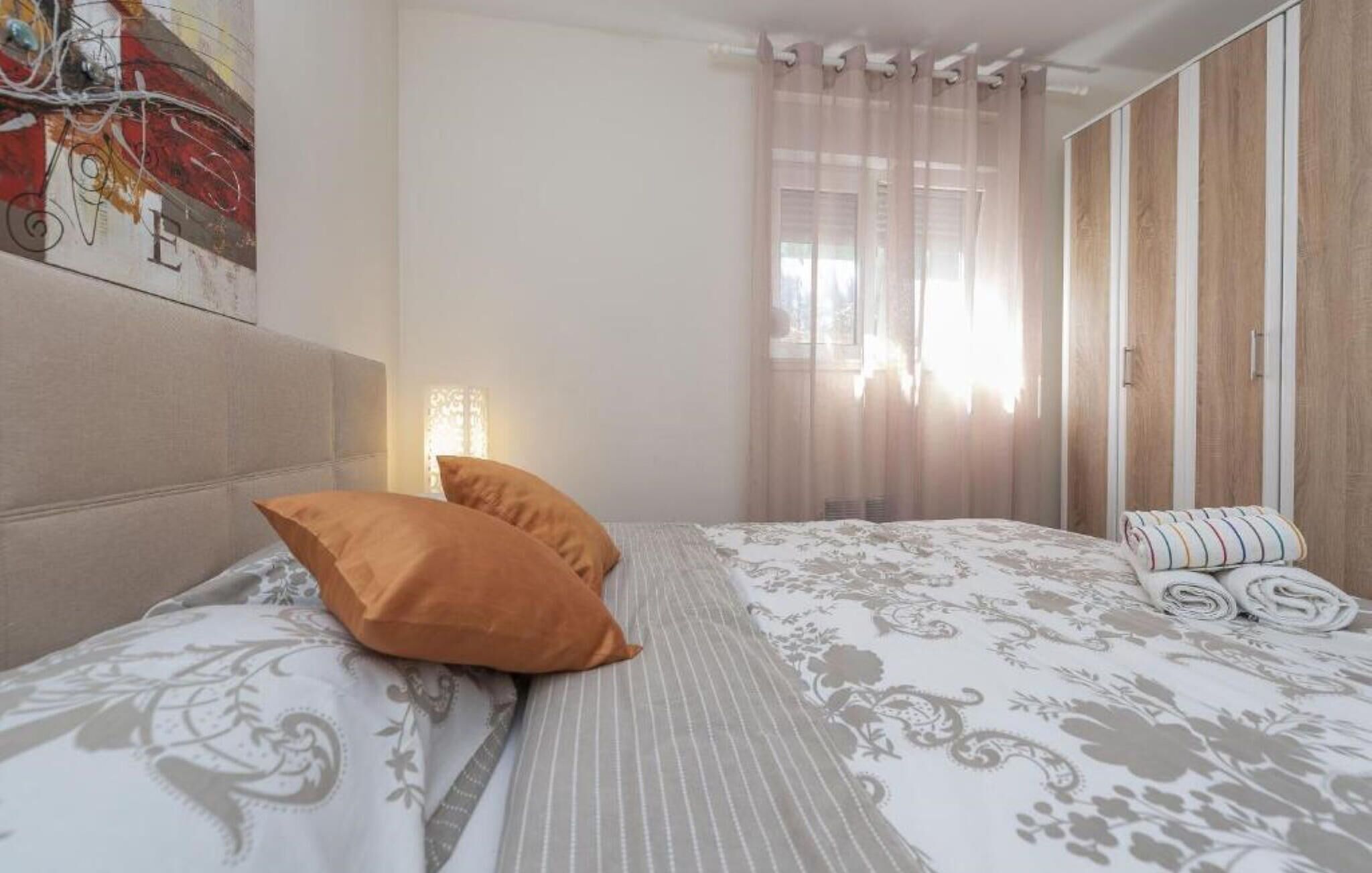 A bedroom with a bed, pillows, a headboard, a window with sheer curtains, and a wooden wardrobe.