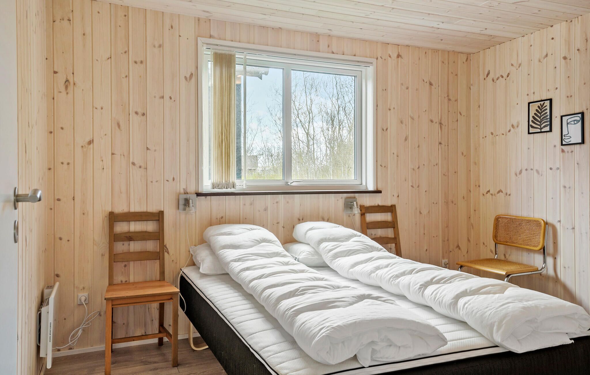 A room with a bed, two wooden chairs, a window, and a wall-mounted shelf.