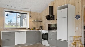 Una cocina moderna con armarios grises, un lavavajillas blanco, una estufa y un refrigerador. Hay una ventana con vistas al exterior, un techo de madera y un taburete de madera.
