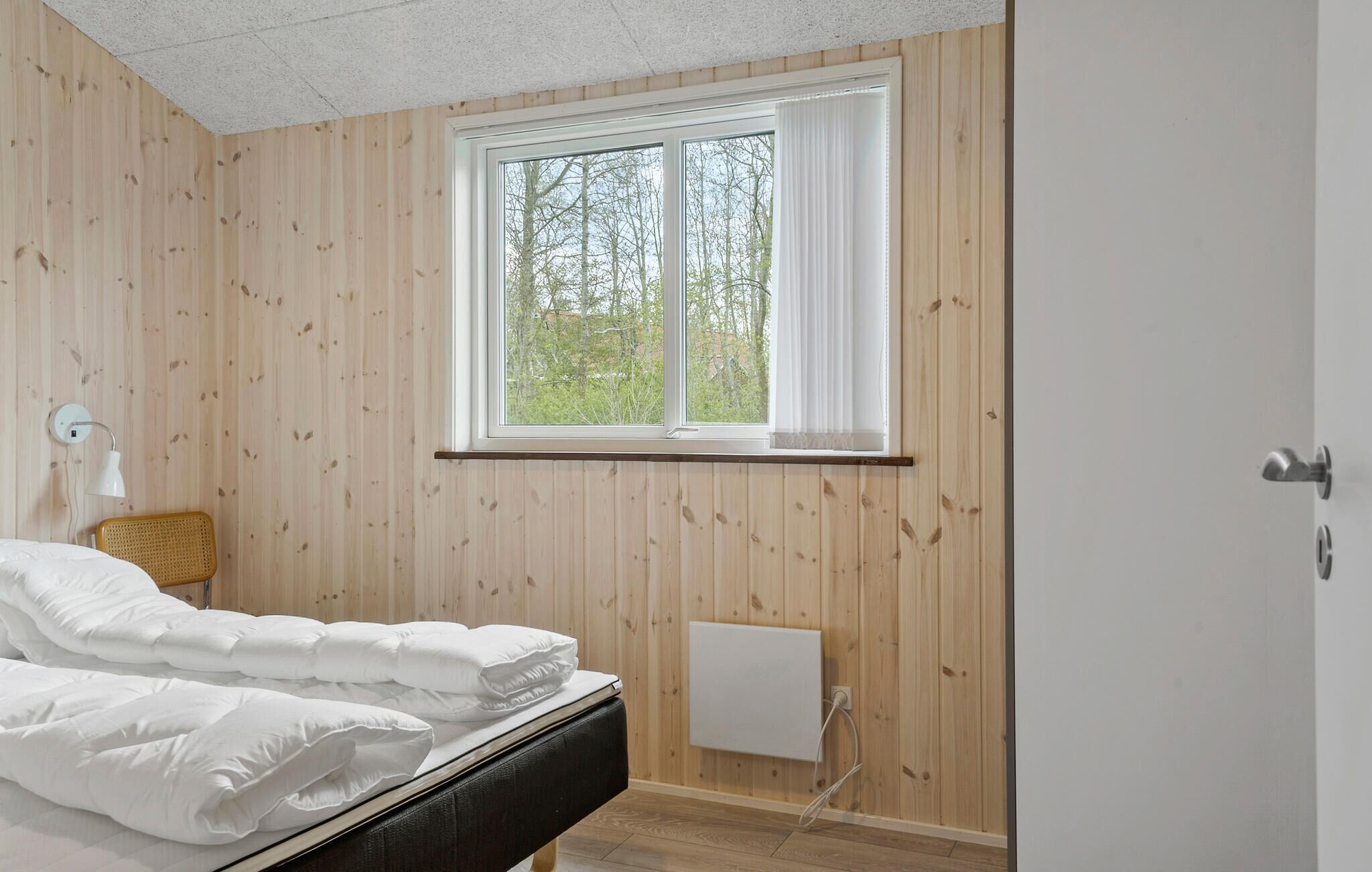 Una camera da letto con pareti in legno, un letto con biancheria bianca, una finestra con tende bianche e una lampada da comodino.