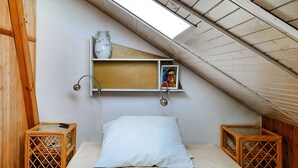 A small, cozy attic bedroom with a sloped ceiling, a single bed, two wooden nightstands, a wall-mounted shelf with a vase and a picture, and a skylight.