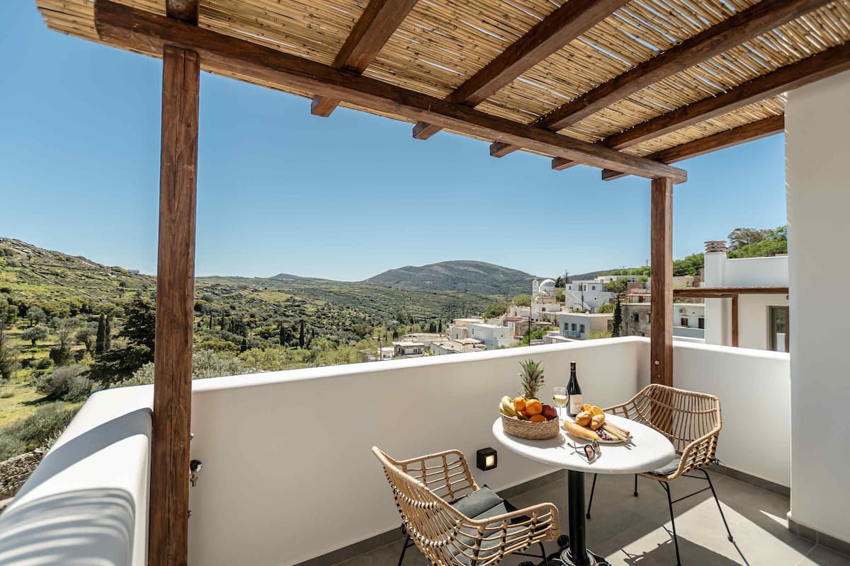 Uma varanda com pérgola de madeira, cadeiras de vime, uma mesa redonda com frutas e uma garrafa, e uma vista panorâmica de colinas e edifícios.