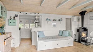 Una habitación acogedora con cama blanca, piso de madera, banco empotrado, pequeña zona de cocina y estufa de leña.