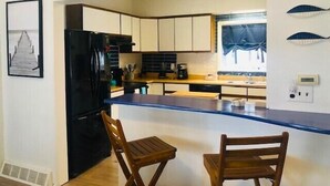 Cocina compacta con armarios blancos, un refrigerador negro y una mesa de comedor de madera con dos sillas. Un letrero sobre la cocina dice "BEACH".