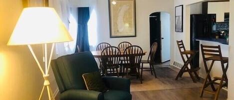 Een woonkamer met een groene bank, een eettafel met stoelen, een plafondventilator en een keuken met een bordje waarop 'BEACH' staat.