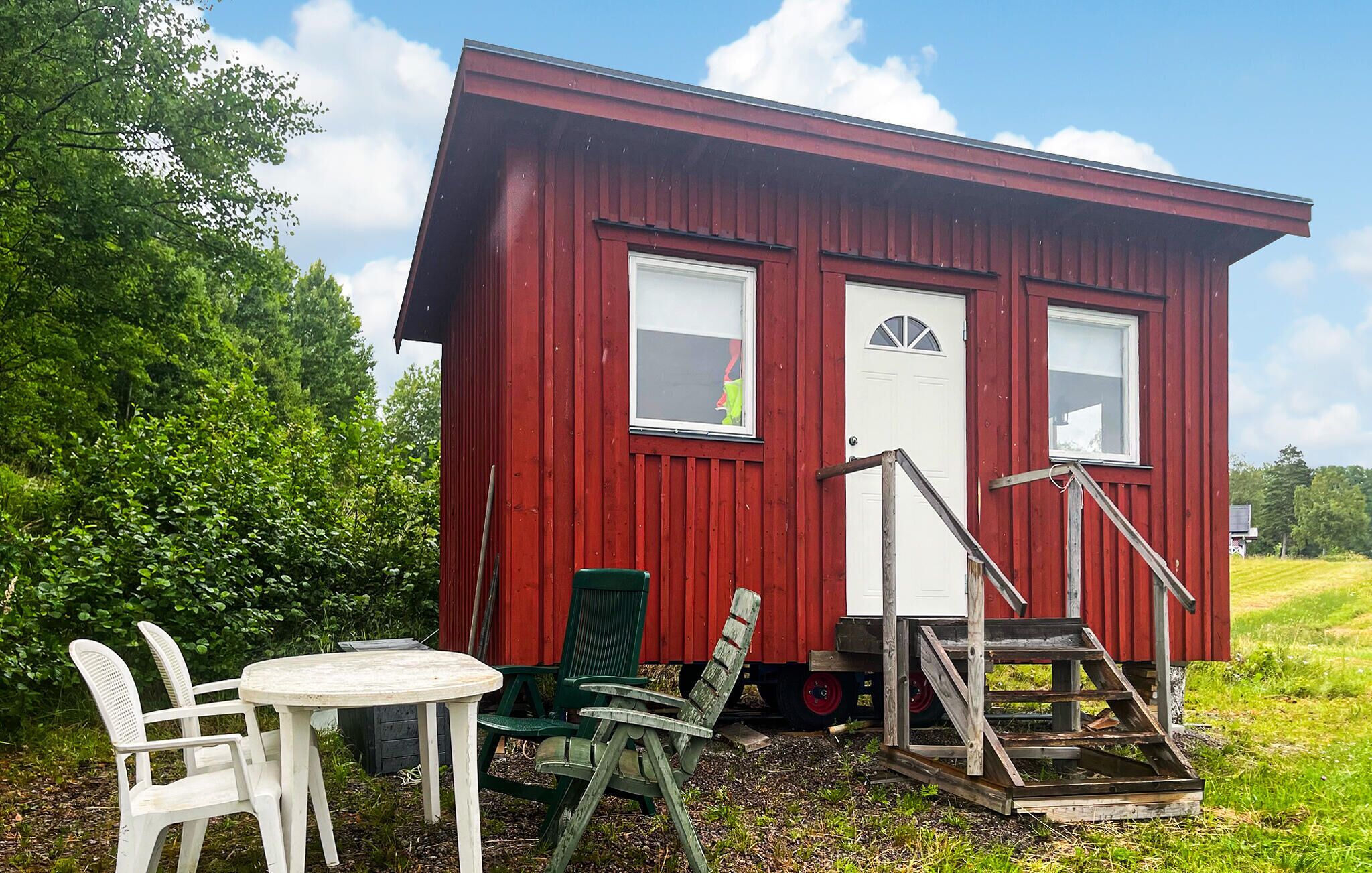 En röd träbyggnad med två fönster och en vit dörr, omgiven av grönska.