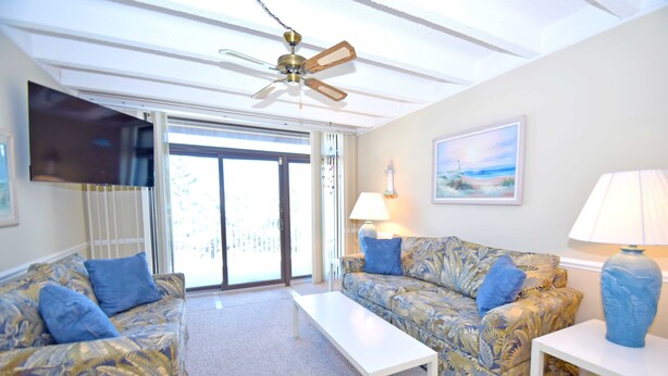 A living room with two patterned sofas, a white coffee table, a ceiling fan, a TV mounted on the wall, and a painting. - Jada 1F-Oceanside 77th St, Free WiFi, Pool, Elev, W/D, AC (Ocean City)