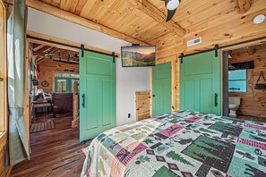 A cozy cabin interior with a bed, green sliding doors, a mounted TV, and a rustic wooden ceiling.