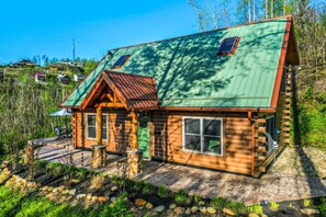 A log cabin with a green metal roof, a stone porch, and a view of a forested area.