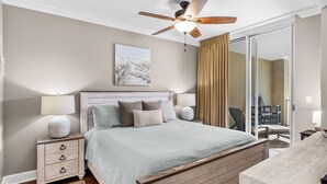 Un dormitorio con cama de madera, lámparas de noche, ventilador de techo, un ventanal con cortinas y un cuadro en la pared.