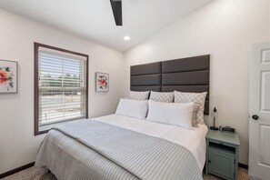 Een slaapkamer met een groot bed, een nachtkastje, een raam met jaloezieën en een deur.